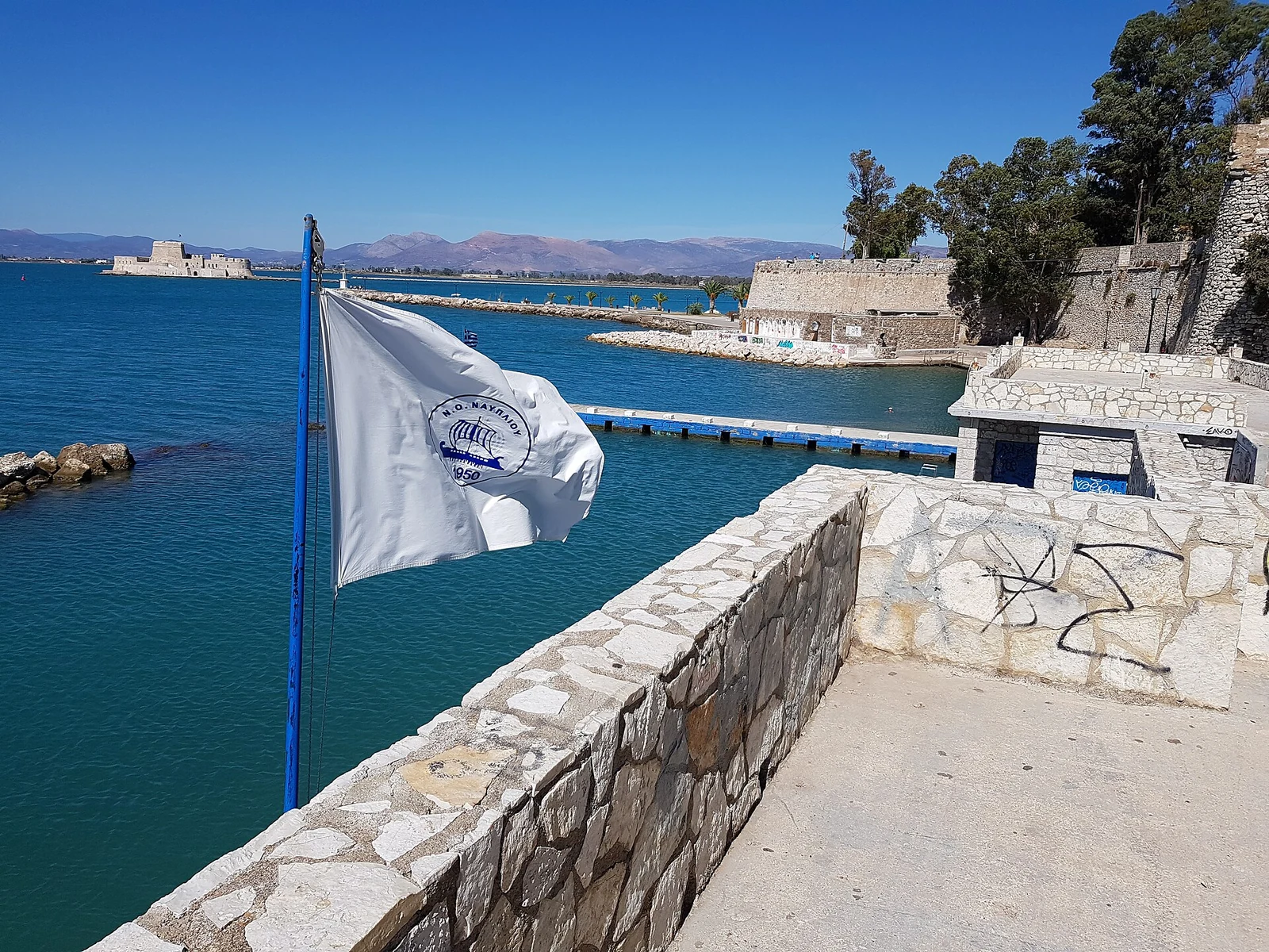 Kamienne platformy nad wodą w Banieres w Nafplio, z flagą na pierwszym planie i fortem Bourtzi w oddali