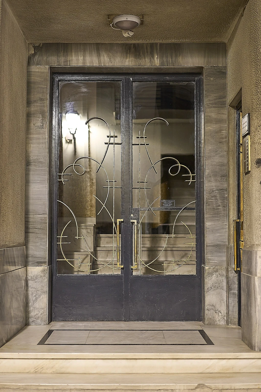 Przeszklone drzwi wejściowe budynku w Plaka w Atenach, z metalowymi ornamentami i schodami za szybą